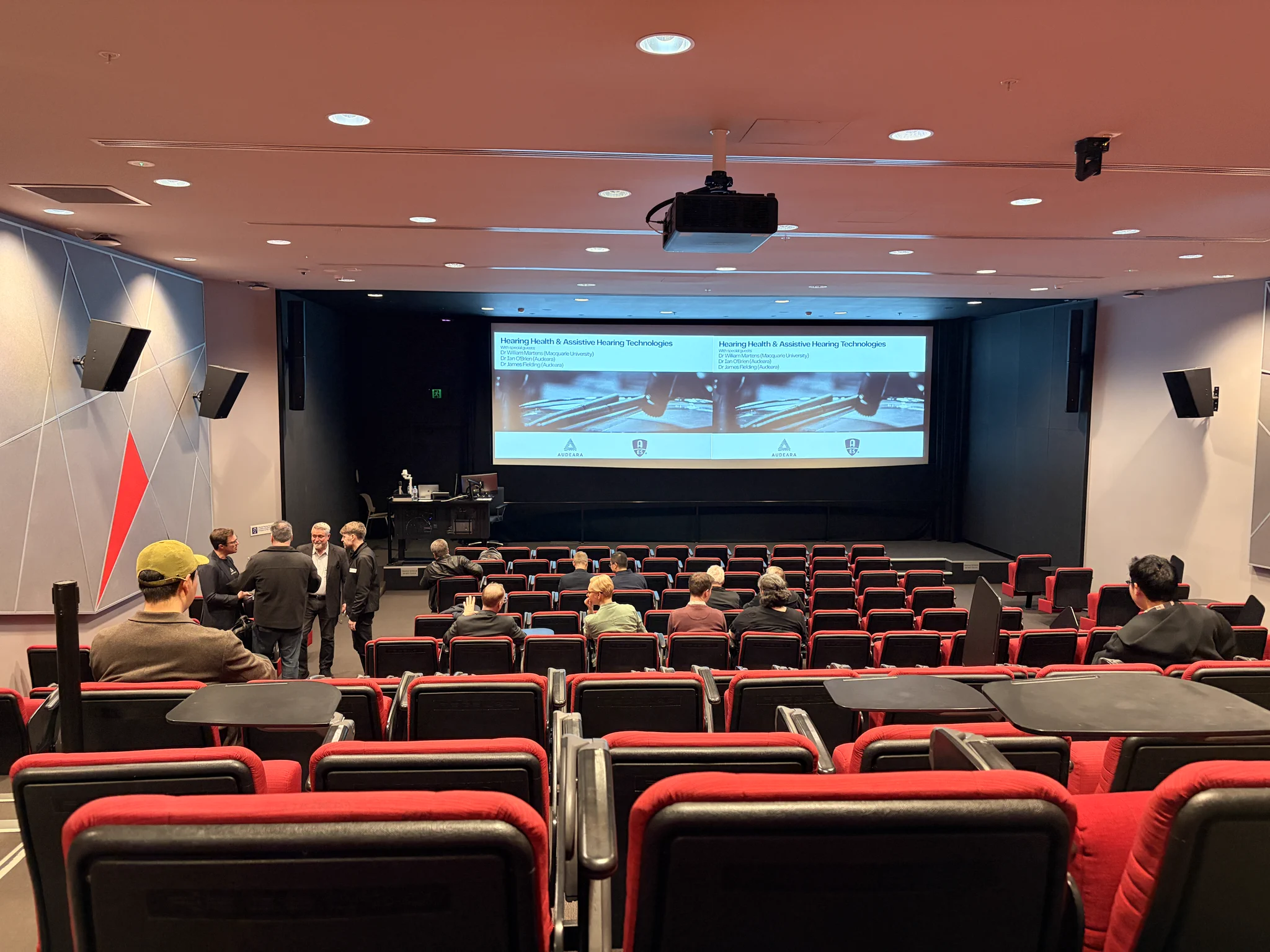 Attendees gather in a lecture theater for an AES Sydney Section meeting on hearing health and assistive hearing technologies, with the event title and speakers displayed on a large screen at the front of the room.