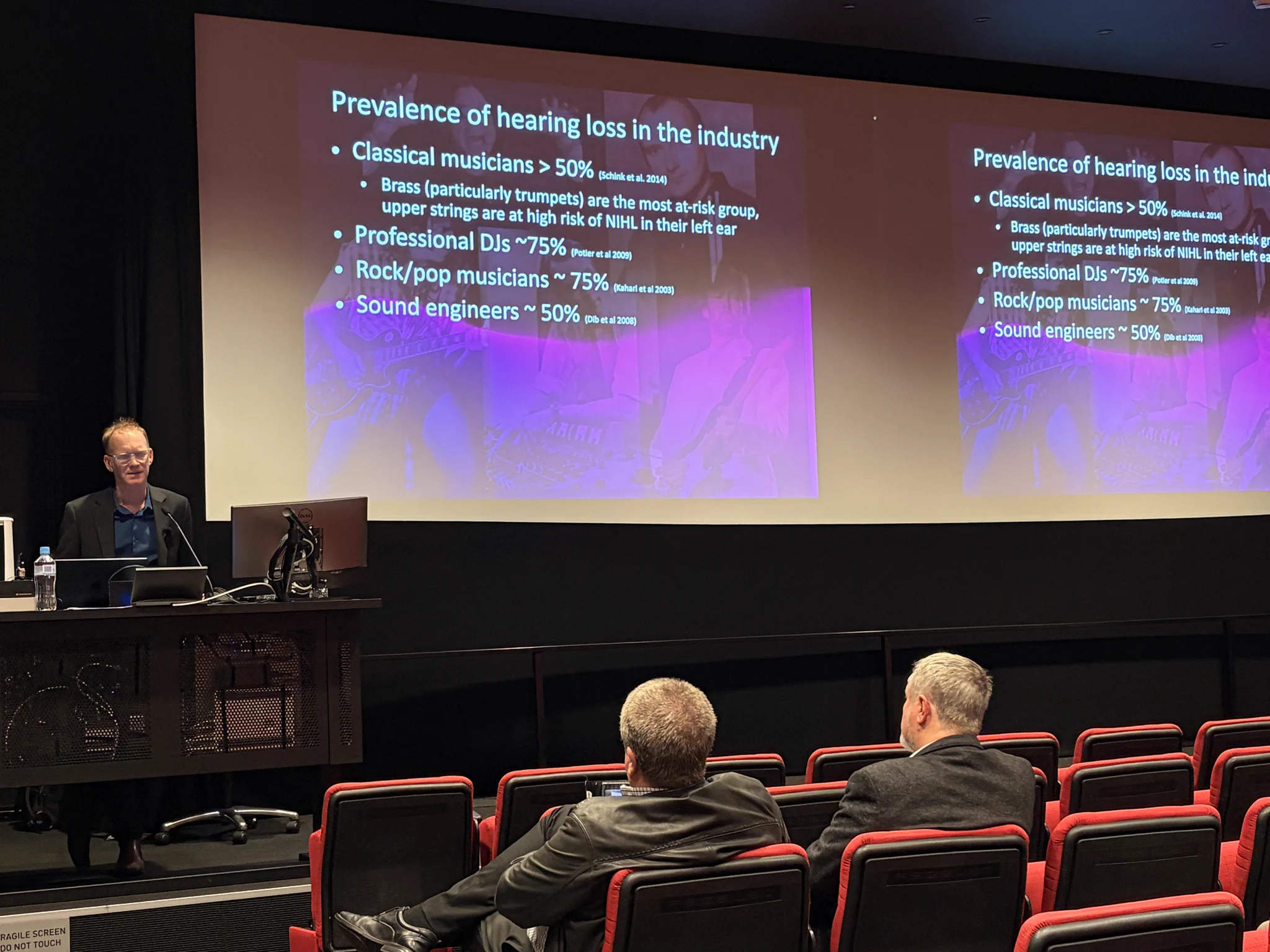 A presenter speaks to AES Sydney Section attendees about the prevalence of hearing loss among musicians, DJs and sound engineers, with supporting statistics displayed on a large presentation screen.
