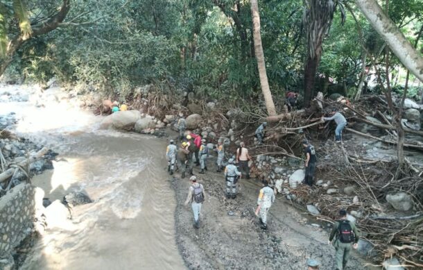An aerial view of a muddy river area in a forest where several people are walking through the debris and water, possibly conducting an investigation in a remote location.