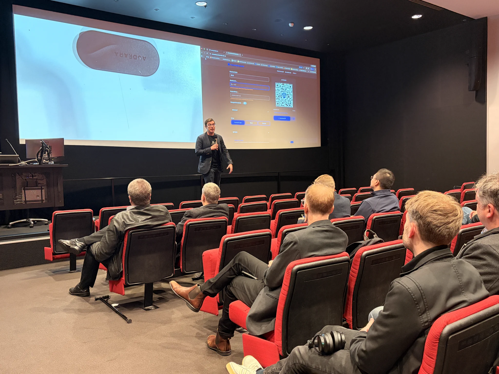 A presenter demonstrates Audeara hearing technology for AES Sydney Section attendees, with a large screen showing the device and its setup interface.