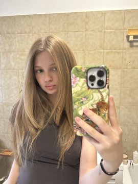 A mirror selfie of a young woman with long blonde hair wearing a brown top, holding a smartphone with a floral patterned case in a tiled bathroom.