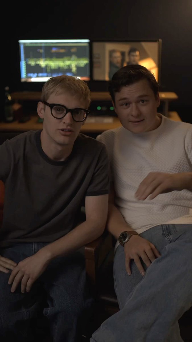 Two young men sit together in what appears to be a dimly lit room featuring dual monitors displaying audio editing software and a movie poster in the background, with the caption suggesting this is a promotional shot for Curry Barker and Cooper Tomlinson's horror comedy film "Anything But Ghosts," releasing in theaters May 7, 2027.