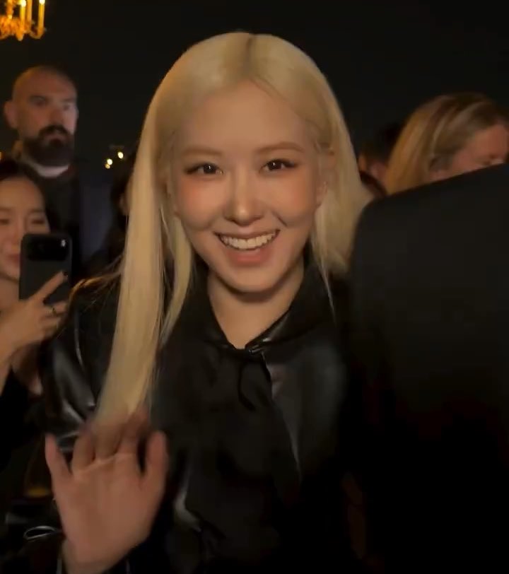 A blonde woman in a black satin blouse smiling and waving at the camera at what appears to be an event, with other people visible in the background.
