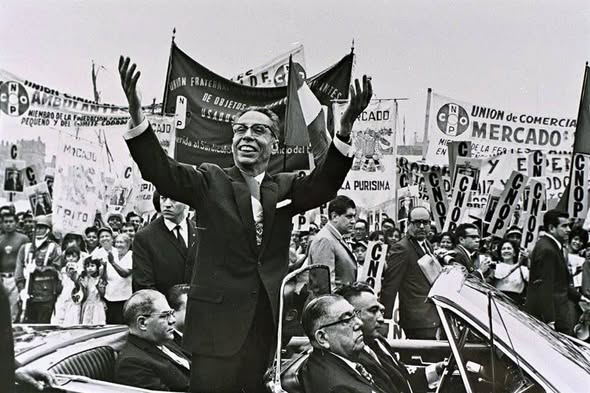 A black-and-white historical photograph of a man in a suit waving enthusiastically from a convertible car, surrounded by a crowd holding banners that read 'UNION DE COMERCIANTES MERCADO.'