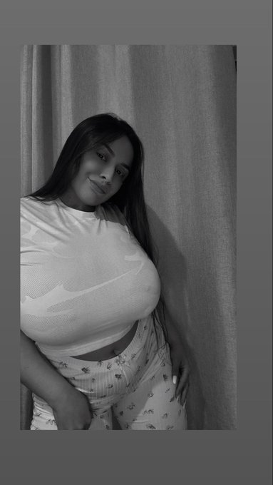 A black and white photo shows a young woman with long dark hair wearing a white Nike crop top and patterned pants, standing against a curtain backdrop.