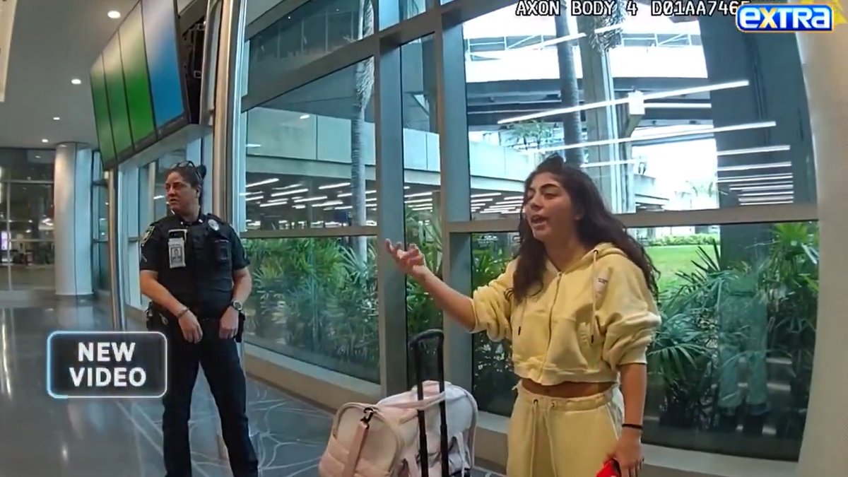 Body camera footage captures a young woman in a cream-colored tracksuit gesturing animatedly while speaking to a female police officer at an airport, with a pink pet carrier visible nearby. The "Extra" news overlay labels the clip "NEW VIDEO" and the top-right timestamp reads "AXON BODY 4 D01AA746," corresponding to Milania Giudice's reported airport outburst.