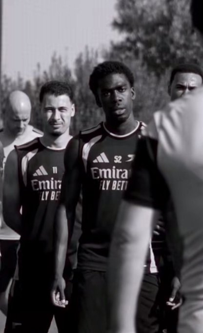 A black and white image showing several soccer players in Arsenal training kits with 'Emirates FLY BETTER' sponsorship, walking together on a training pitch.