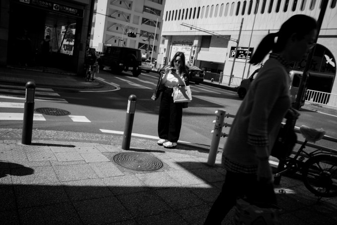 A black-and-white street photograph captures two women at an urban intersection in Japan, with one woman in the foreground walking past and the other crossing the street in the background.