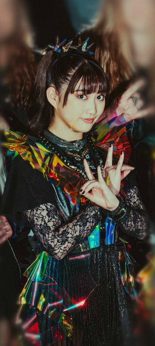 A young woman with dark hair and bangs poses making a peace sign, wearing an elaborate iridescent black costume with spiked accessories and lace sleeves.