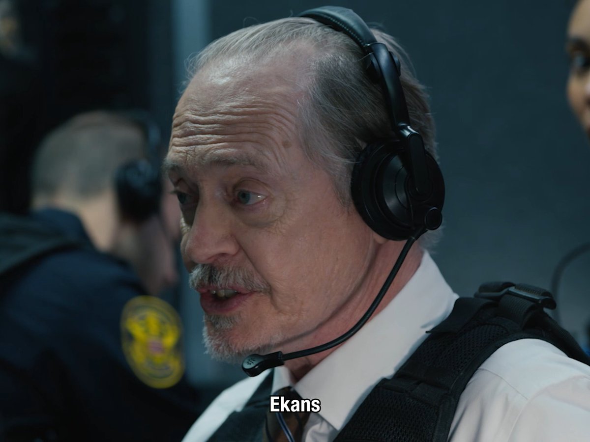A caption describing what is in the image: A middle-aged man with gray hair, mustache, and headset with microphone, wearing a white shirt and black tie with a tactical vest, speaks intensely while the subtitle "Ekans" appears at the bottom of the screen. He appears to be in a law enforcement or tactical setting, with another officer partially visible behind him, and the word "Ekans"—a deep-cut reference to the Pokémon snake character—serves as a punchline to whatever tense scenario is unfolding in the scene from "The Terrors of Jordan Mendoza."