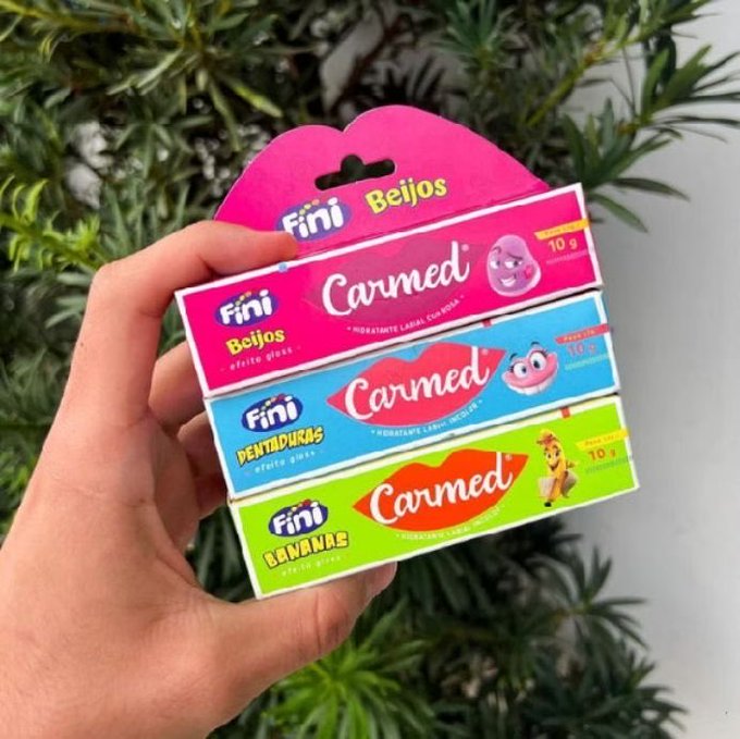 A hand holds a pack of three Fini brand Carmed lip glosses in Beijos (pink), Dentaduras (blue), and Bananas (green) flavors, each 10g.