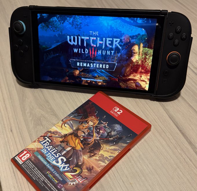 A Nintendo Switch 2 displaying 'The Witcher 3: Wild Hunt Remastered' on screen, with a physical copy of 'Trails in the Sky 2nd' game case placed below it on a light wooden table.