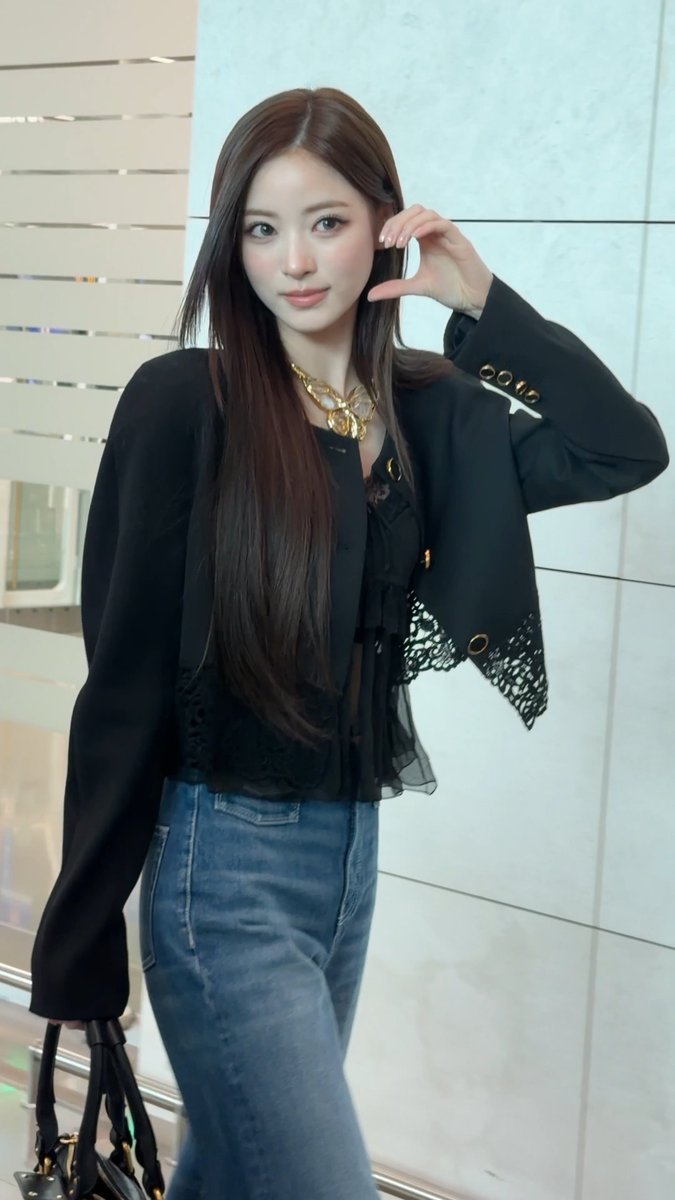 A young woman with long dark hair poses indoors wearing a black lace-trimmed blazer over a sheer blouse, blue jeans, and a chunky gold chain necklace.