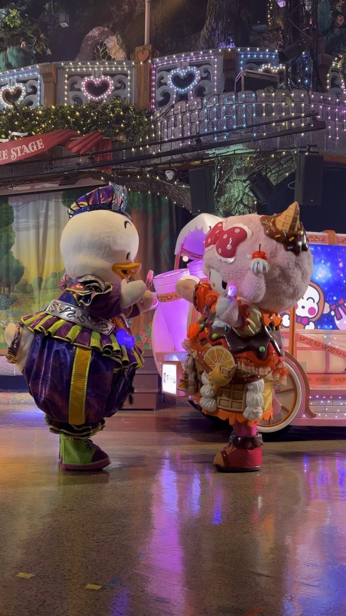 A photograph of two costumed character mascots — a duck in a purple jester outfit and a pink dessert-themed character — performing on a brightly lit stage with 'SANRIO' illuminated in the background.