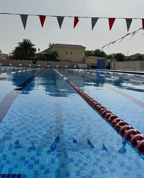 An empty outdoor swimming pool with lane dividers and backstroke flags strung overhead, set against a clear sky with a residential building in the background.
