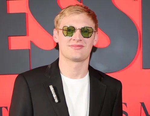 A man with reddish-blonde hair wearing green-tinted aviator sunglasses, a white t-shirt, and a black blazer with a decorative pin, standing in front of a red and black backdrop.