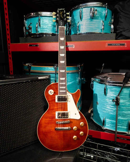 An Epiphone Les Paul Standard 50s electric guitar with a Root Beer finish is in front of a set of turquoise drum kits on a red shelf. An amplifier and microphone are visible nearby.