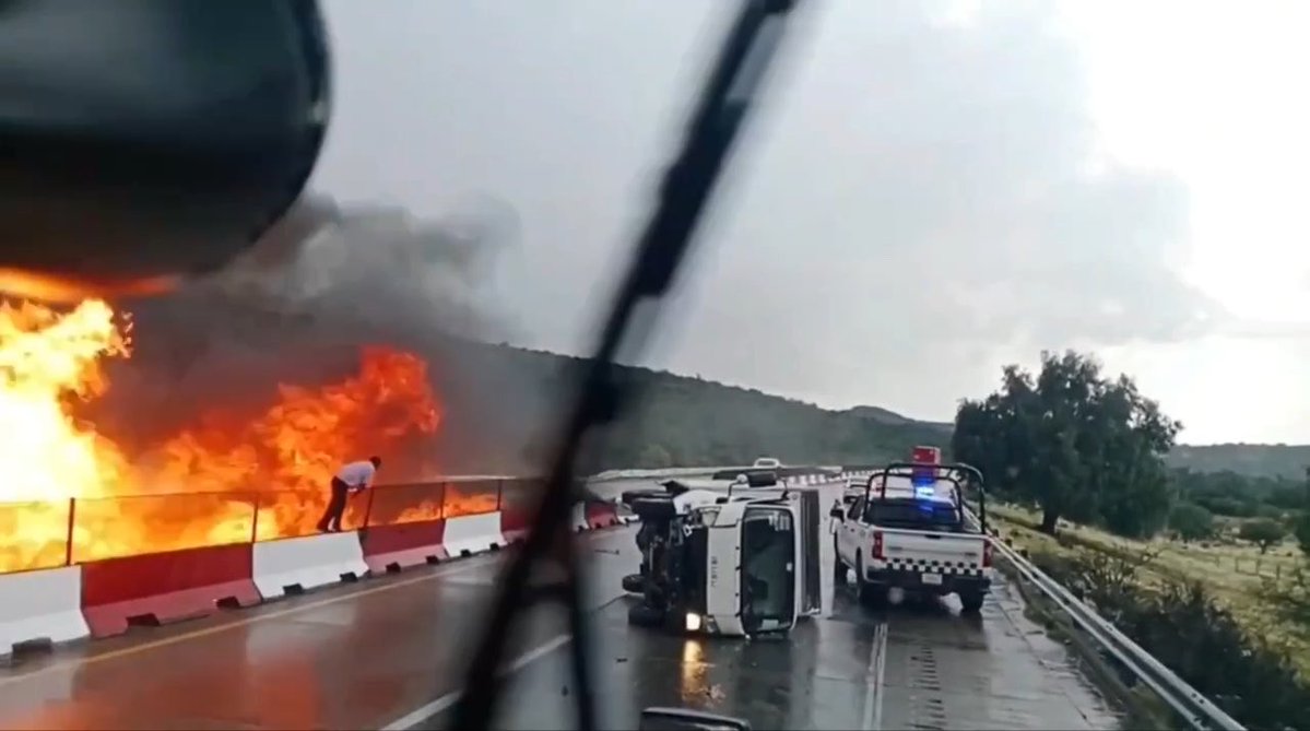 A man runs from a massive fire on a Mexican highway after a crane crashes and erupts into flames, with an overturned crane and a police pickup truck marked with emergency lights visible at the scene. The image captures the dramatic moment of the man's narrow escape from the blazing wreckage, aligning with the post's description of the fiery accident and his seconds-long dash to safety.