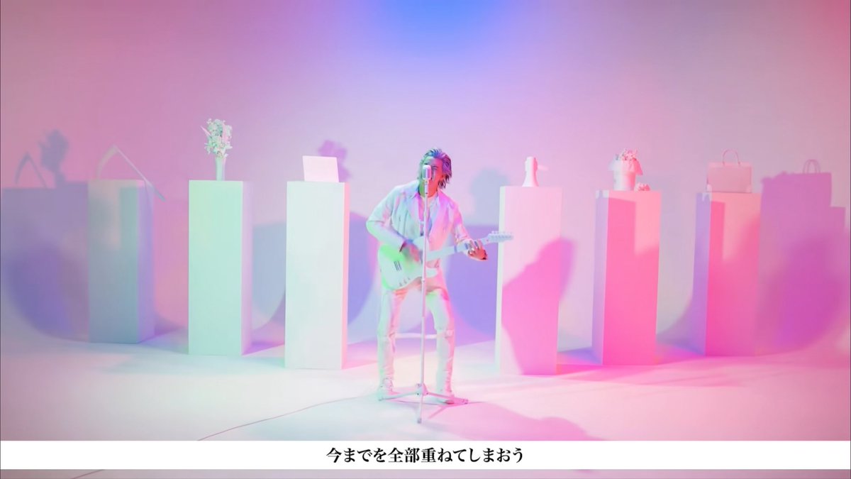 A musician plays guitar on a pastel-colored stage surrounded by pedestals with flowers and accessories, with Japanese subtitle '今までを全部重ねてしまおう' at the bottom.