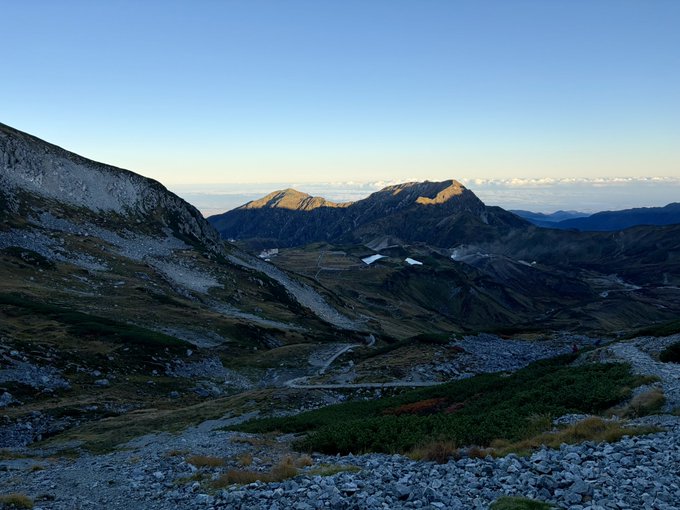 A scenic mountain landscape at what appears to be sunrise or sunset, with a winding trail visible in the rocky terrain and a small lake in the distance.