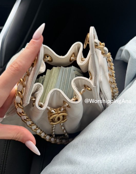 A photo of a hand holding open a white quilted Chanel drawstring bag filled with stacks of cash, with the watermark '@WorshippingAna' visible.