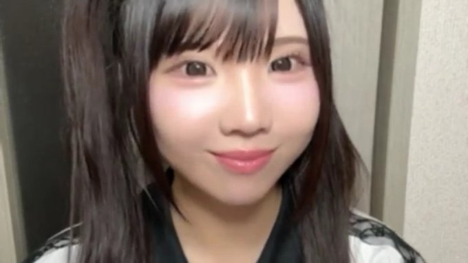 A close-up portrait of a young woman with long dark hair and bangs, wearing pink lipstick and a white top with a black collar.