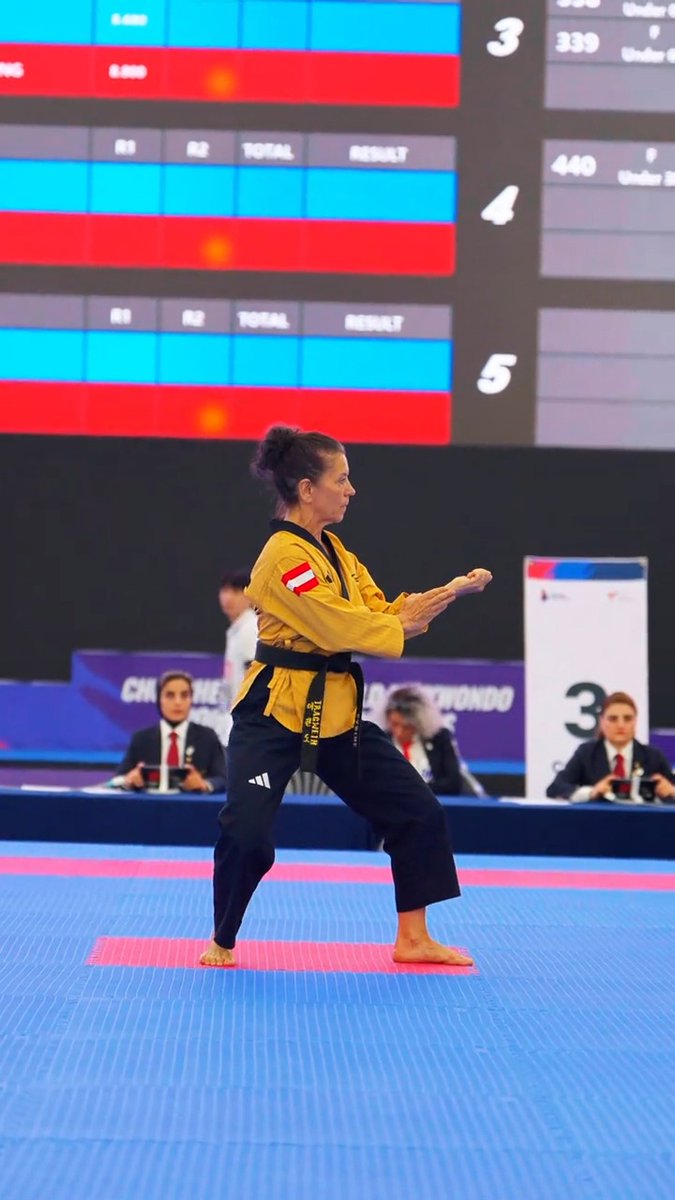 A female martial artist in a yellow and black uniform performs a stance on a blue and red mat during a championship competition, with a digital scoreboard visible in the background.