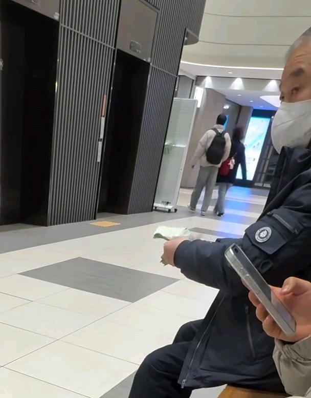 A masked older man sits on a station bench holding up Japanese yen bills to display, while another person in the foreground takes the photo with their phone. The scene captures the exact moment referenced in the caption, where the man is ostentatiously showing off his cash in a public transit area.