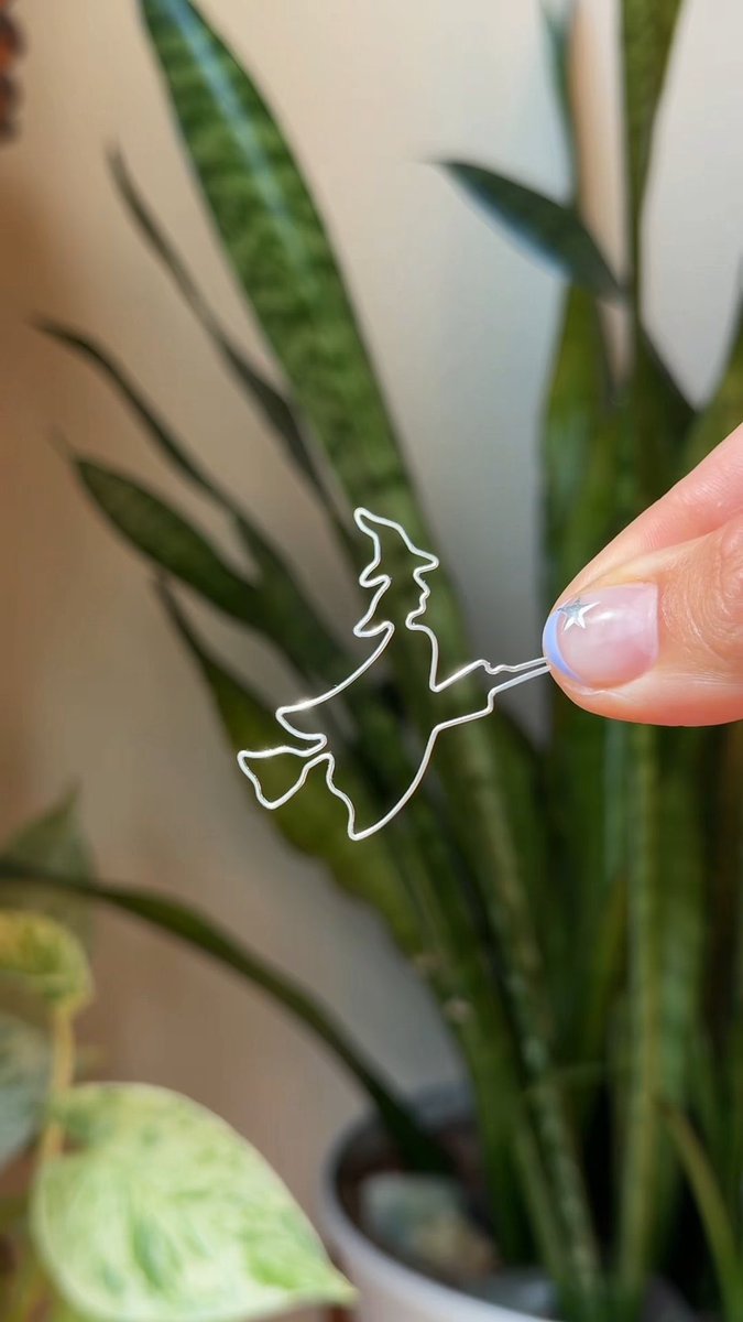 A hand with manicured nails holding a small clear acrylic cutout shaped like a witch on a broomstick, with a snake plant in the background.