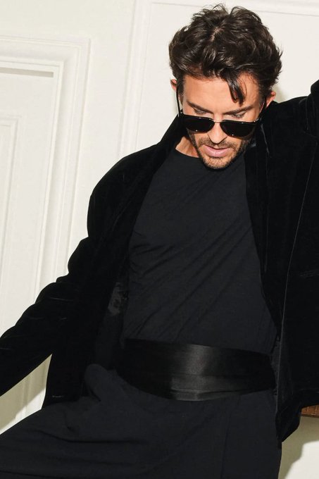 Jonathan Bailey poses in a dramatic all-black ensemble featuring a tailored coat, crew neck top, and sleek belt for L'Officiel Hommes, channeling a brooding, masculine elegance as he looks downward through dark sunglasses.