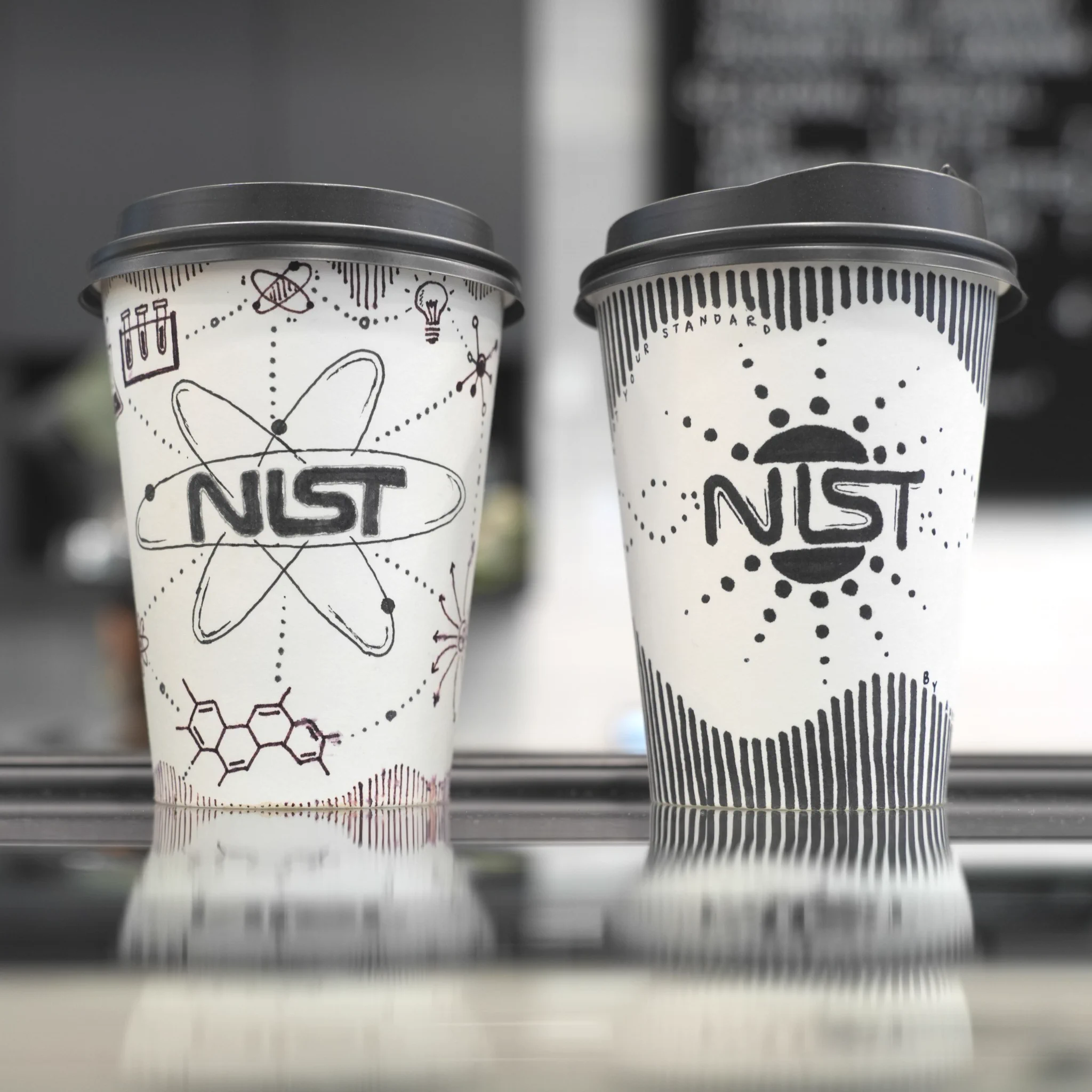 Two coffee cups on a reflective surface, each featuring the NIST logo. The cups have scientific-themed designs, including atomic symbols and molecular structures. A blurred blackboard is in the background.
