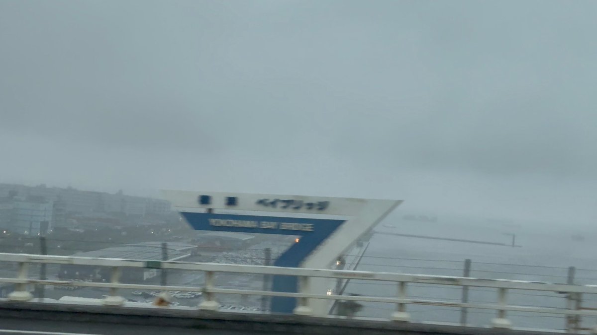 Foggy view of a port or bay with a Japanese sign that appears to read "KAIKAN" (横浜港シンボルタワー), taken from a bridge or elevated road with railings in the foreground.