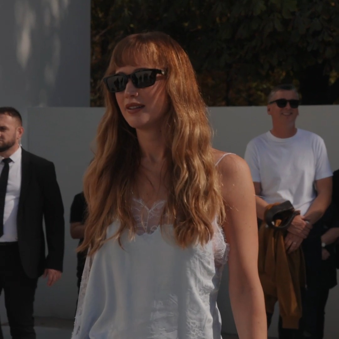 A woman with long red hair and bangs wears black sunglasses and a white lace dress while walking outdoors, with a security guard and onlookers visible in the background.