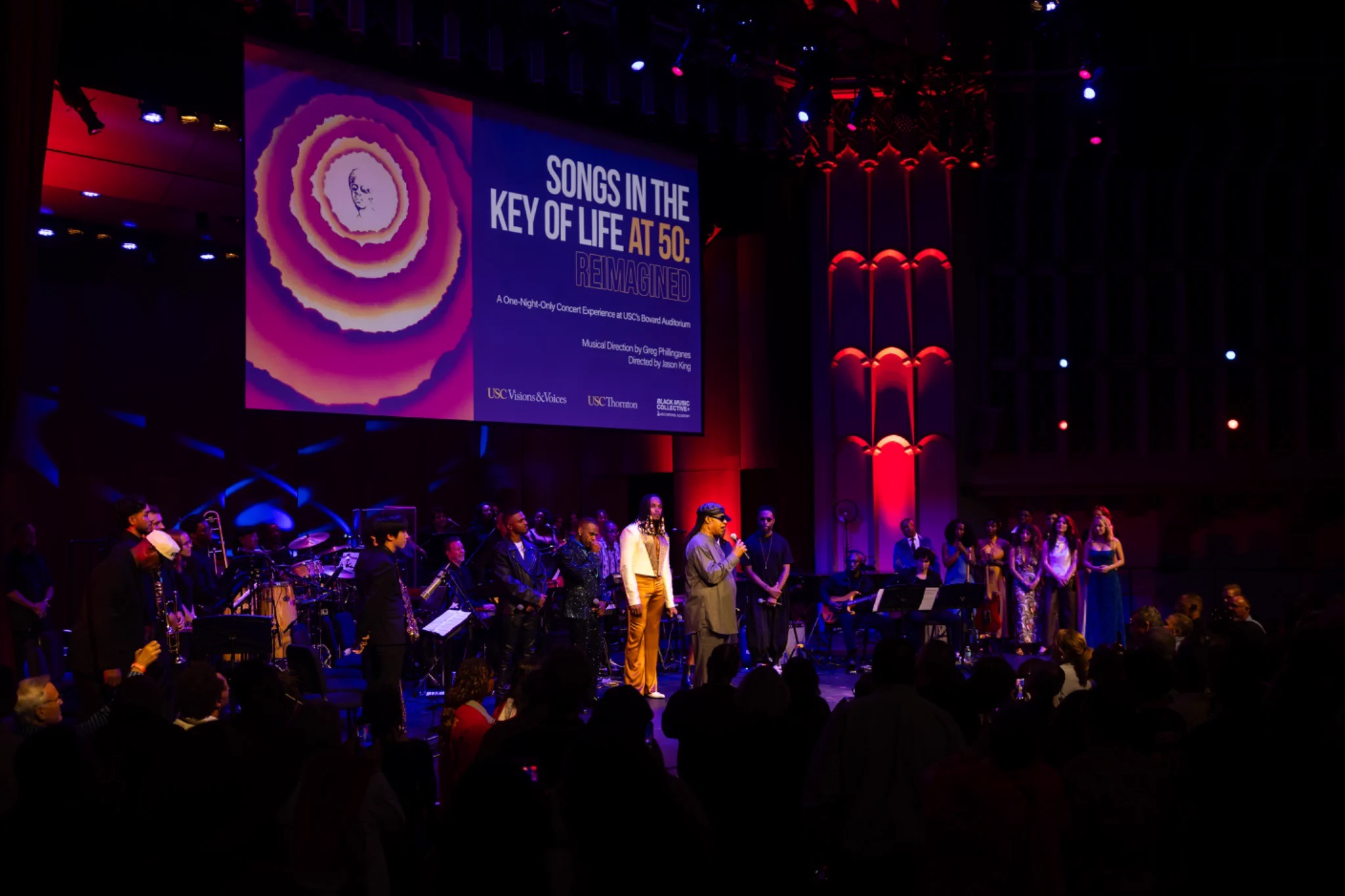 A performer speaks into a microphone onstage surrounded by musicians and singers as an audience watches. A large screen above reads “Songs in the Key of Life at 50: Reimagined.”