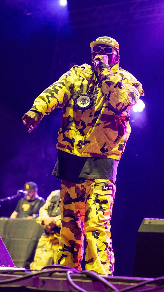 A performer in a yellow camouflage Supreme outfit and cap, wearing sunglasses and a large chain, holds a microphone on a purple-lit stage, with a second person in matching camo gear visible in the background. The image connects to the post by visually representing Public Enemy's iconic stage presence, complementing the caption about Flavor Flav and Chuck D as rap pioneers promoting social justice through music.