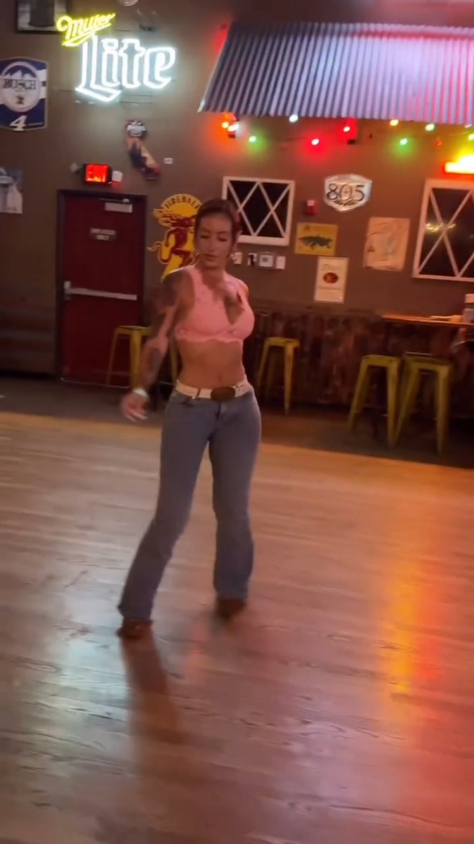 A woman in a pink top and jeans performs a line dance on the wooden floor of "Miller Lite" bar, with visible signage including "HCH 4," "Supernatural," and "805," likely captured mid-spin to emphasize the "don't blink" warning.