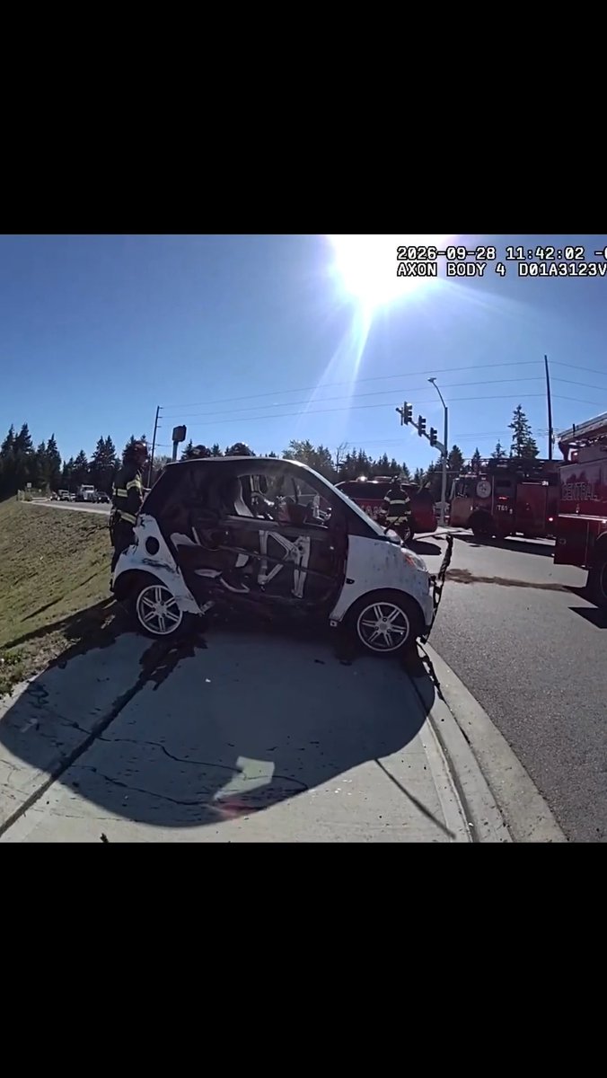 Body camera footage timestamped 2026-09-28 11:42:02 showing the aftermath of a car accident involving a heavily damaged small white car, with firefighters and a fire truck on scene.