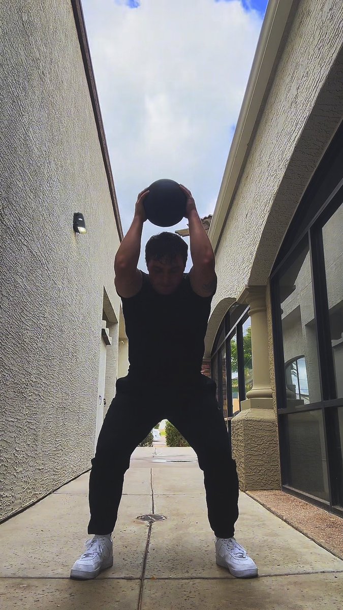 A man in all-black athletic wear and white sneakers performs a squat-style exercise while holding a black medicine ball overhead in a narrow outdoor walkway between two buildings.
