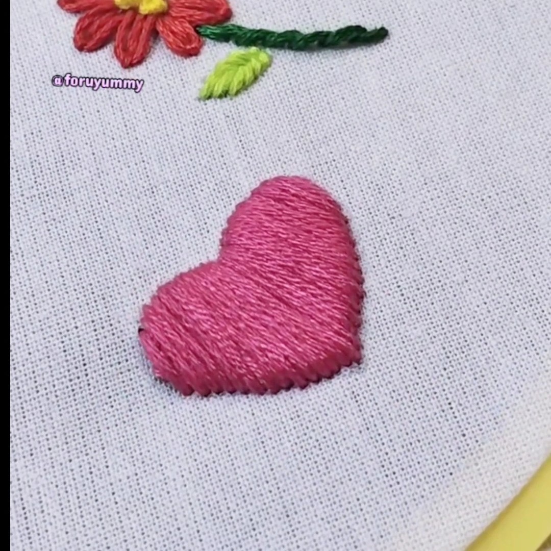 A close-up of embroidery on white fabric showing a pink stitched heart below a red flower with green leaves, watermarked '@foruyummy'.