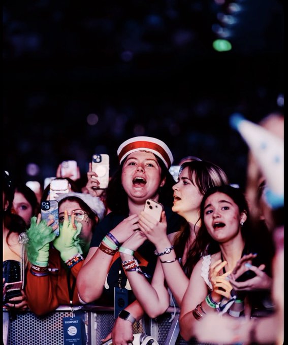 A group of young concertgoers react emotionally at a live event, with one girl in a white cap crying while holding up her phone, surrounded by others with wristbands and phones raised.