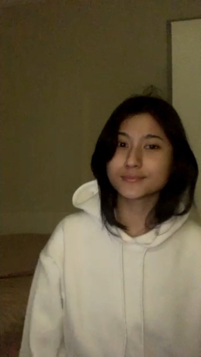 A selfie of a young woman with dark shoulder-length hair wearing a white hoodie, taken indoors against a plain wall.