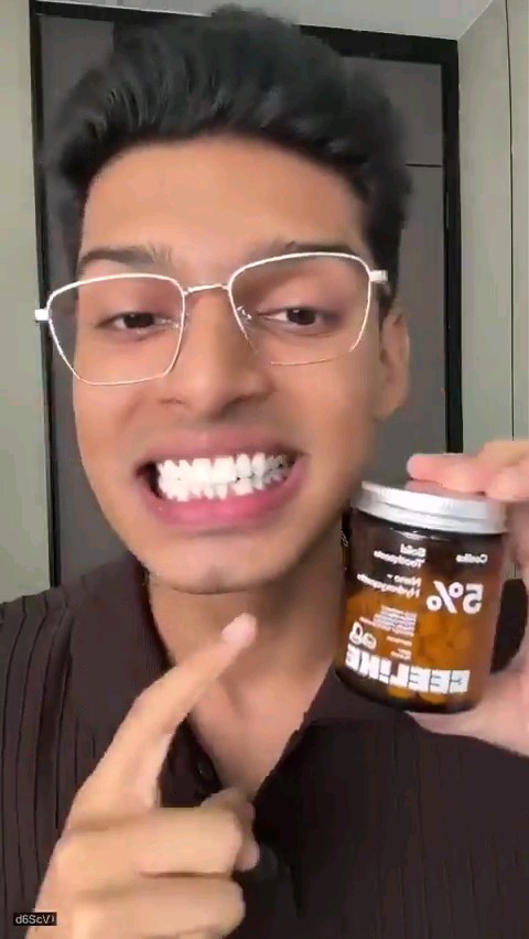 A man with glasses smiles widely, showing prominent diamond-encrusted teeth, while holding a brown beverage can and pointing at it.