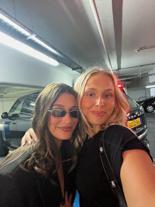 Two women smile for a close-up selfie inside a parking garage, with a car visible in the background.
