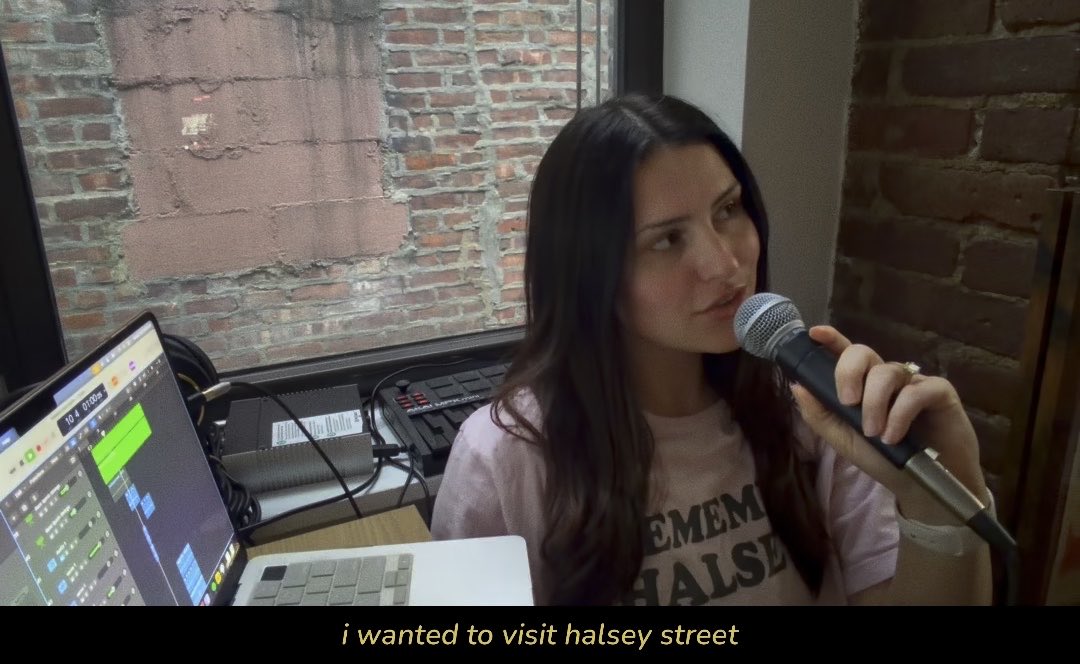 A woman with long dark hair speaks into a microphone while sitting in front of a laptop running music production software, with the caption 'i wanted to visit halsey street' displayed at the bottom.