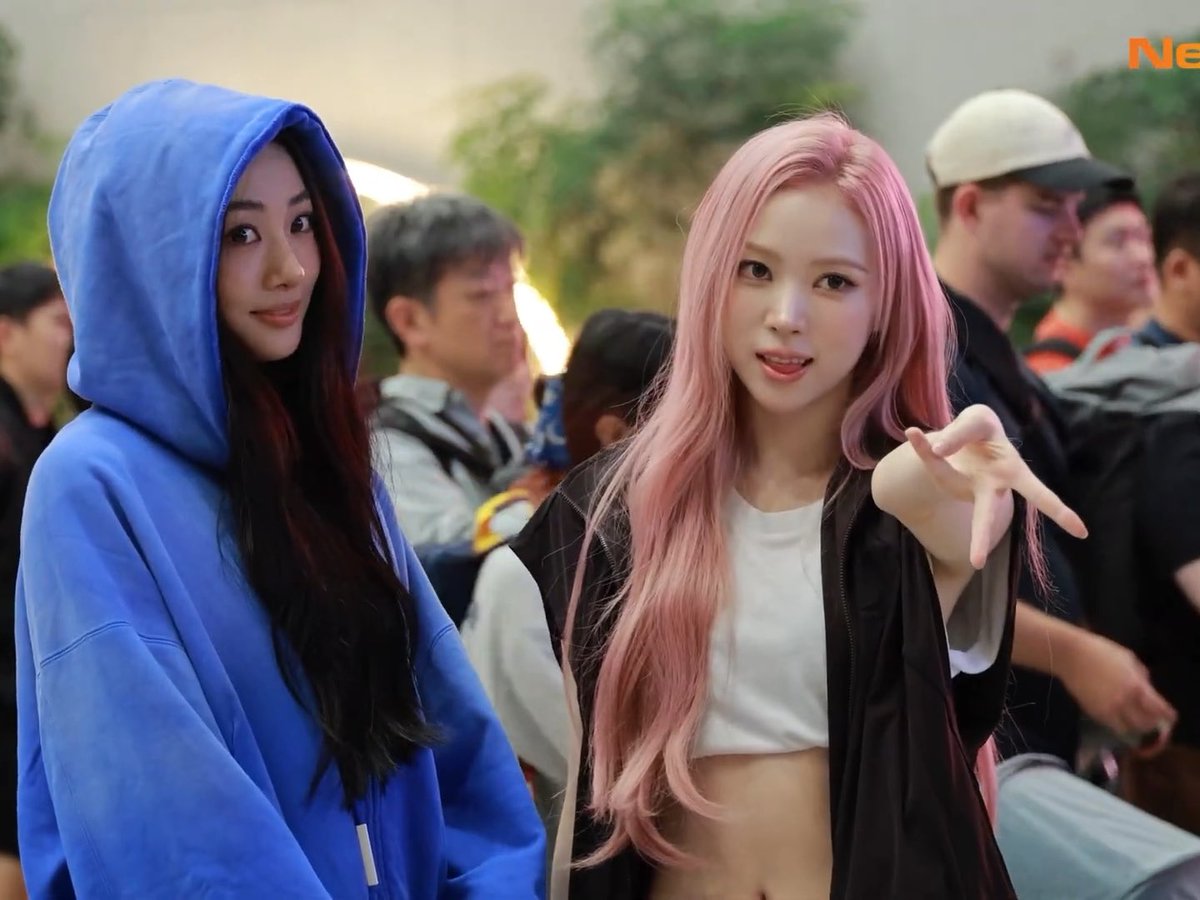 The same two women from the previous image are seen in a closer shot, one in the blue hoodie and the other with pink hair reaching her hand toward the camera, with 'Ne' text visible in the corner.