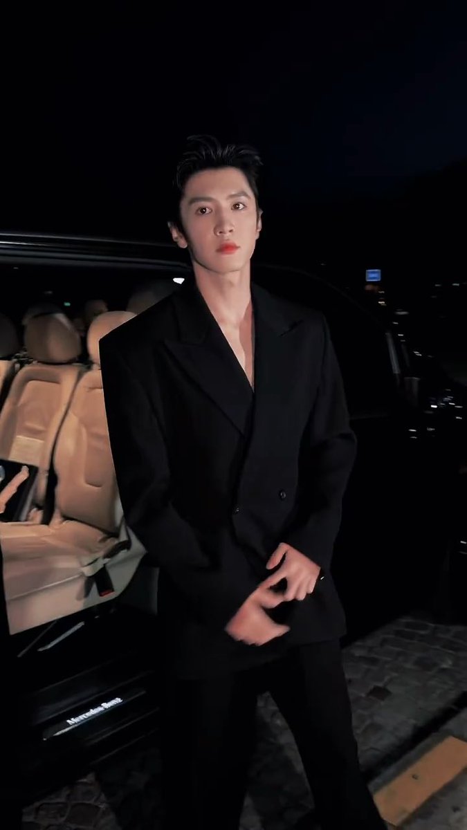 A man in a black double-breasted suit stands at night beside an open luxury vehicle (Mercedes-Benz) with tan leather interior.