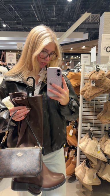A woman with blonde hair and glasses takes a mirror selfie in a shoe store while holding a brown leather boot and a Coach purse, wearing a dark gray jacket.