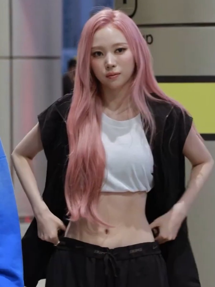 A woman with long pink hair stands with her hands on her hips, wearing a white crop top and a black vest over black sweatpants.