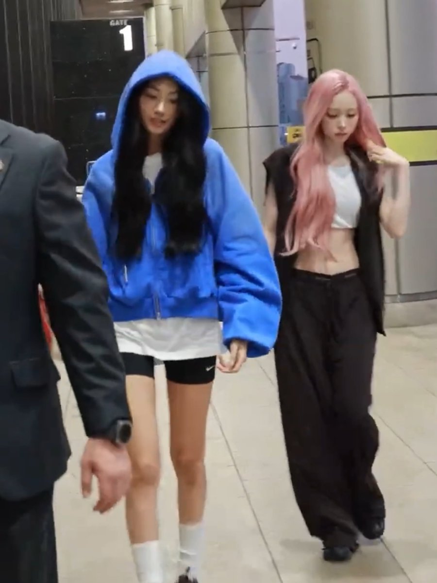 Two women, one wearing a blue hooded jacket and the other with pink hair, walk through what appears to be an airport terminal area marked 'GATE 1'.