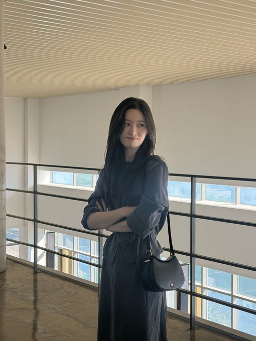 A woman with long dark hair standing indoors in a blue dress, arms crossed, holding a black crescent-shaped handbag, with windows visible behind her.